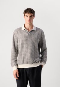 Young man wearing a long-sleeve knit sweater with white collar and cuffs, paired with black pants, standing against plain background.