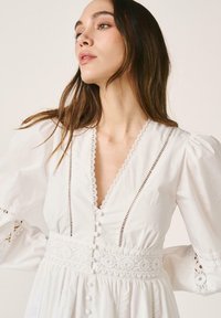 Jeune femme aux longs cheveux bruns portant une robe blanche avec une bordure en dentelle, des manches bouffantes et des boutons décoratifs sur le devant.