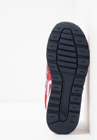 Semelle de chaussure avec un motif de relief profond, semelle extérieure en caoutchouc noir, partie supérieure rouge et blanche visible sur le côté, positionnée verticalement contre un mur de coin blanc.