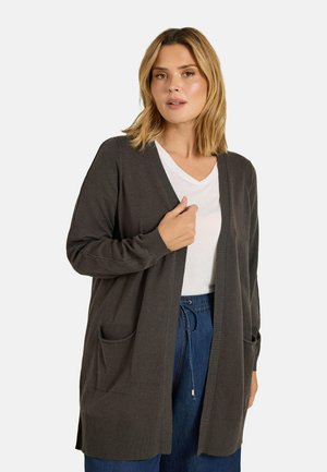 Femme portant un cardigan gris foncé ouvert sur un t-shirt blanc à col en V et un pantalon bleu à cordon, sur un fond clair uni.
