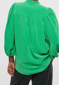Persoon draagt een groene blouse met opgeblazen mouwen, gezien vanachter. De stof heeft een subtiele textuur, gecombineerd met zwarte broek.
