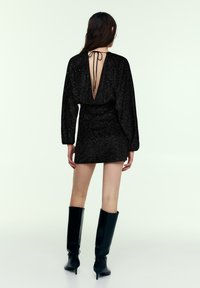 Černé třpytivé mini šaty s dlouhými rukávy, hlubokým výstřihem na zádech do V a zavazováním. Nošeny s černými vysokými kozačkami na podpatku po kolena. Hladká textura a přiléhavý střih.