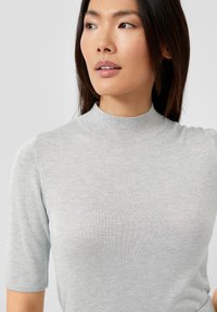 Hellgraues Strickoberteil mit kurzen Ärmeln und hohem Kragen, das ein dezentes strukturiertes Muster und eine weiche, leicht reflektierende Oberfläche aufweist.