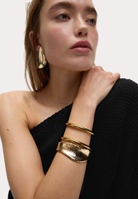 Boucles d'oreilles en métal doré en forme de goutte, bracelets en or superposés, et un bracelet en or texturé. Top en tricot noir laissant l'épaule à découvert.