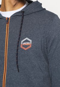 Marinblå zip-hoodie med orange accenter; har en hexagonal logotyp. Tygtexturen är slät med ett subtilt mönster.