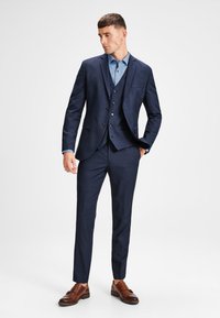 Costume marine ajusté, avec une veste à un bouton, des revers à cran et trois boutons. Associé à une chemise bleu clair et des chaussures marron.