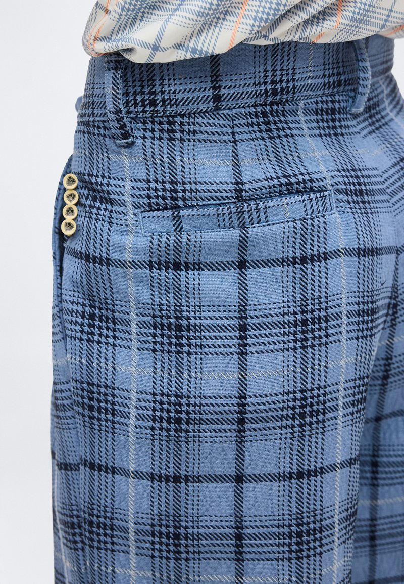 Pantalones a cuadros azules con líneas blancas y negras, que cuentan con un bolsillo y cuatro botones beige en el lateral, combinados con una camisa estampada.