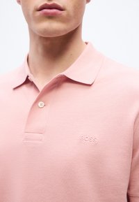 Κοντινή λήψη ενός ατόμου που φοράει ανοιχτό ροζ πόλο μπλουζάκι BOSS με γιακά και ένα κουμπί κουμπωμένο, εμφανίζοντας το κεντημένο λογότυπο.