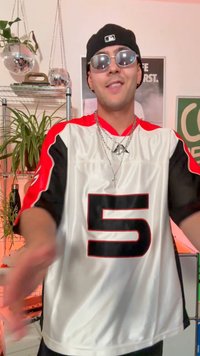 Joven con gafas de sol, gorra de béisbol y camiseta con el número 5, de pie en una habitación con plantas y bolas de disco colgantes.