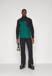 Chaqueta de forro polar en verde oscuro y negro, con cierre frontal y un bolsillo en el pecho. Combinada con pantalones de esquí negros y botas de esquí, sosteniendo equipo de esquí.