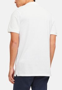 Polo shirt bianco a maniche corte con colletto, spacchi laterali all'orlo e texture liscia. Cuciture visibili e etichetta del marchio sul lato.
