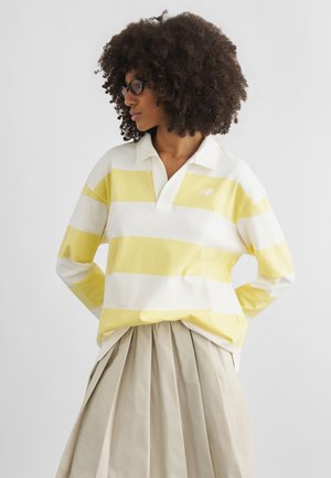 Woman with curly hair and glasses wearing a yellow and white striped rugby shirt and beige pleated skirt, looking to her left.