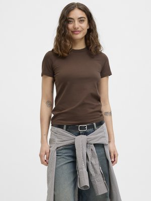 Ung kvinde med bølget brunt hår iført en brun T-shirt, blå jeans og en stribet skjorte bundet omkring taljen, smiler blidt.