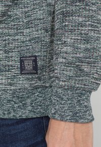 Suéter verde y gris con tejido texturizado, con un dobladillo ajustado y una pequeña etiqueta de logo que muestra "Scotch & Soda" y "Amsterdam Couture."