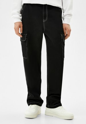 Homme portant un pantalon cargo noir avec des coutures blanches et des baskets blanches, debout sur un fond blanc uni.