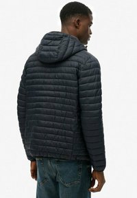 Mand iført en sort quiltet hættejakke og blå jeans, stående mod en ensfarvet hvid baggrund, med ryggen til kameraet.