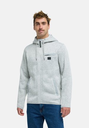 Mężczyzna w jasnoszarej kurtce z kapturem na suwak z kieszeniami i niebieskich dżinsach, stojący na tle gładkiej białej ściany.