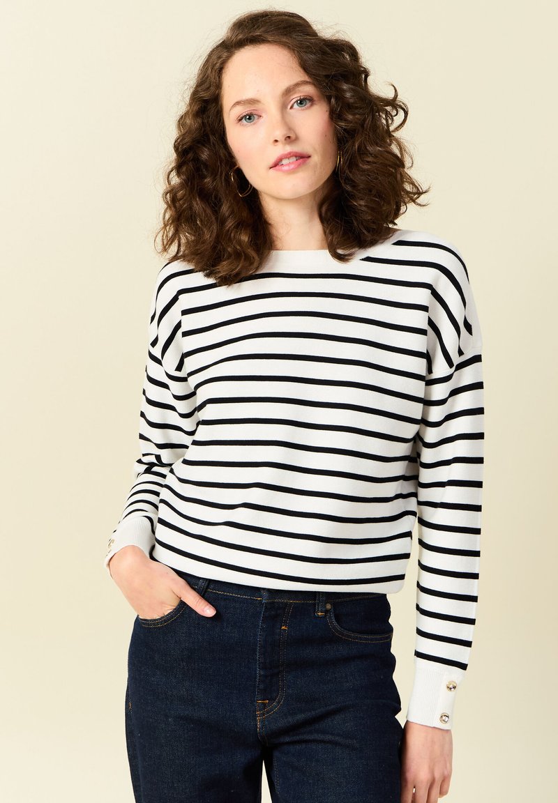 Suéter de rayas en blanco con líneas horizontales negras, con escote barco y mangas acampanadas con detalles de botones. Combinado con jeans oscuros.