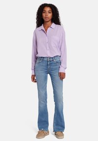 Chemise longue à manches longues violette claire avec des boutons et une poche, associée à un jean bleu clair évasé et des chaussures beiges à enfiler.