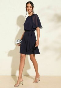 Námornícka modrá šaty s voľnou chiffon kápou, s nariaseným výstrihom a detailom opasku viazaného v páse. Skombinované so striebornými sandálmi na opätku s remienkami.