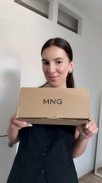 Rjava kartonska škatla z natisnjenim "MNG" v črni barvi. Škatlo drži oseba v črnem oblačilu, v svetlem notranjem prostoru.