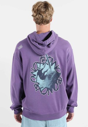 Personne aux cheveux courts portant un sweat à capuche violet affichant des graphismes de surfeur en bleu et noir ainsi qu'un texte au dos.