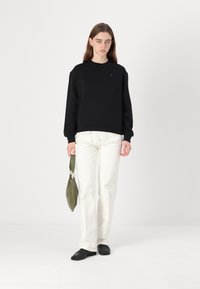 Felpa nera con colletto rotondo, maniche lunghe e dettaglio del logo sul petto, abbinata a jeans bianchi a gamba dritta e una borsa verde.