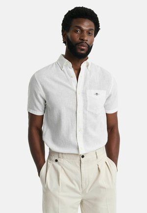 Short-sleeve white linen shirt with a pointed collar, front pocket, and subtle embroidery detail. Paired with light beige trousers.