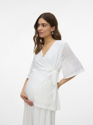 Femme enceinte en robe portefeuille blanche à festons avec larges manches, tenant son ventre de la main gauche, tournée vers la gauche sur un fond uni.