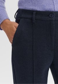 Ruka v kapse tmavých strukturovaných kalhot, které jsou doplněny světle fialovým pleteným svetrem, na kterém jsou vidět detaily knoflíku a poutka na opasek.