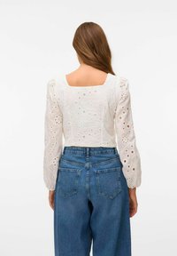 Bílý vyšívaný crop top s dlouhými, naddýmavými rukávy, s květinovými vzory a výřezy. Kombinován s vysoce pasovými modrými džíny.