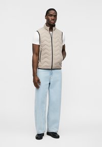 Άνδρας που φοράει μπεζ βαμβακερό γιλέκο πάνω από λευκό T-shirt με ανοιχτό μπλε τζιν παντελόνι με φαρδιά μπατζάκια και μαύρα παπούτσια, στέκεται μπροστά σε απλό φωτεινό φόντο.