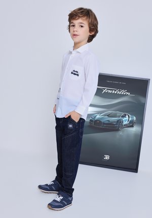 Un ragazzo giovane con camicia bianca e jeans scuri è accanto a un poster incorniciato di un'auto sportiva blu Bugatti Chiron con la scritta "Tourbillon".