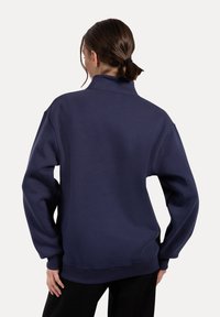 Persona con el cabello oscuro recogido lleva un sudadera holgada de color azul marino, de pie con las manos cerca de las caderas, de espaldas en un fondo liso.