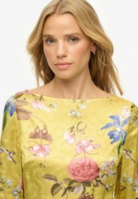 Geel bloemenjurk met een satijnen afwerking, versierd met roze rozen en blauwe orchideeën. Het ontwerp heeft een brede halslijn en lange mouwen.