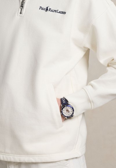 Άτομο που φοράει λευκό πουλόβερ Polo Ralph Lauren με φερμουάρ, με το χέρι στην τσέπη, και μπλε ρολόι Polo Ralph Lauren στον καρπό.