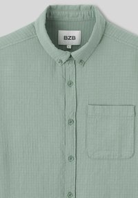 Chemise boutonnée vert clair en tissu texturé. Dotée d'un col, de cinq boutons et d'une poche sur la poitrine gauche.
