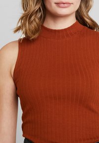 Kvinna iförd ärmlös, ribbad, bränd orange topp med hög halsringning, som visar en del av ansiktet och axellångt vågigt hår.