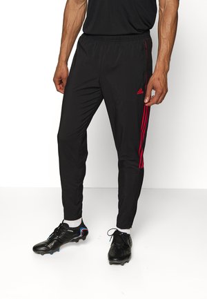 Mężczyzna ubrany w czarne spodnie dresowe Adidas z czerwonymi paskami oraz czarne korki piłkarskie z białymi skarpetami, stojący na białym tle.