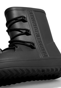 Botín de goma negro con superficie texturizada, diseño con cordones y detalles de logo en relieve a lo largo del costado. Presenta una suela de tracción robusta.