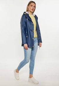 Marinblå vattentät jacka med huva, gula knappar och sidofickor; matchad med en randig gul och vit topp, ljusblå jeans och vita sneakers.