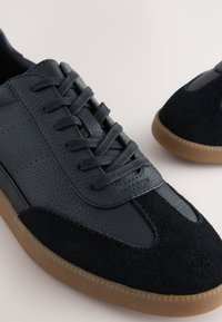 Črni supergi iz usnja in semiša z teksturirano površino, okroglim noskom in zavezovanjem z vezalkami. Podplat iz rjavega gume.