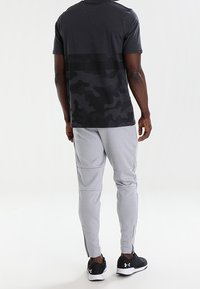 Conjunto deportivo masculino que incluye una camiseta negra con patrón de camuflaje y pantalones jogger grises ajustados con cremalleras en los tobillos, combinados con zapatillas negras.