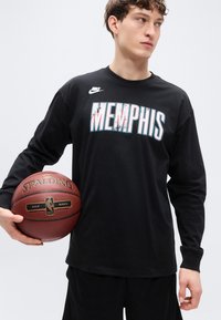 Μαύρο T-shirt μακρυμάνικο με το γραφικό «MEMPHIS», το λογότυπο της Nike και μια μπάλα του μπάσκετ στο χέρι. Κανονική εφαρμογή με ριμπ μανσέτες, φορεμένο με μαύρο σορτς.
