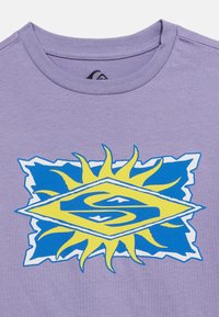 Μωβ μπλουζάκι με γραφικό εκρηκτικό ηλιακό μοτίβο σε μπλε και κίτρινο χρώμα και ένα στυλιζαρισμένο λογότυπο "S" στο κέντρο του στήθους κάτω από τον γιακά.