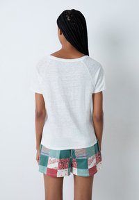 Camiseta blanca, ligera, con cuello redondo y mangas cortas, combinada con pantalones cortos de patchwork coloridos que presentan una mezcla de patrones y texturas.