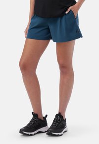 Turkosa träningsshorts med en slät textur, elastisk midja och sidofickor. Bärs med svarta löparskor med vita sulor.