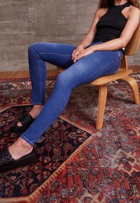 Vaqueros skinny azul oscuro con un diseño clásico de cinco bolsillos, combinados con zapatos negros de tejido sin cordones, sentado en una silla de madera.