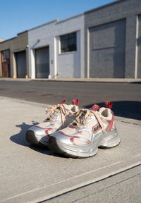 Grove zilveren en rode sneakers met beige veters geplaatst op een zonovergoten stoep voor industriële gebouwen met garagedeuren.
