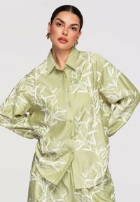 Femme portant une chemise boutonnée vert clair et un ensemble pantalon avec un motif de feuilles blanches, debout les mains sur les hanches devant un fond uni.